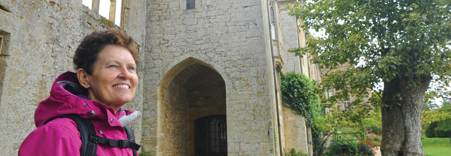 A smiling woman wearing a backpack and pink jacket stands before a stone castle in England or Wales.