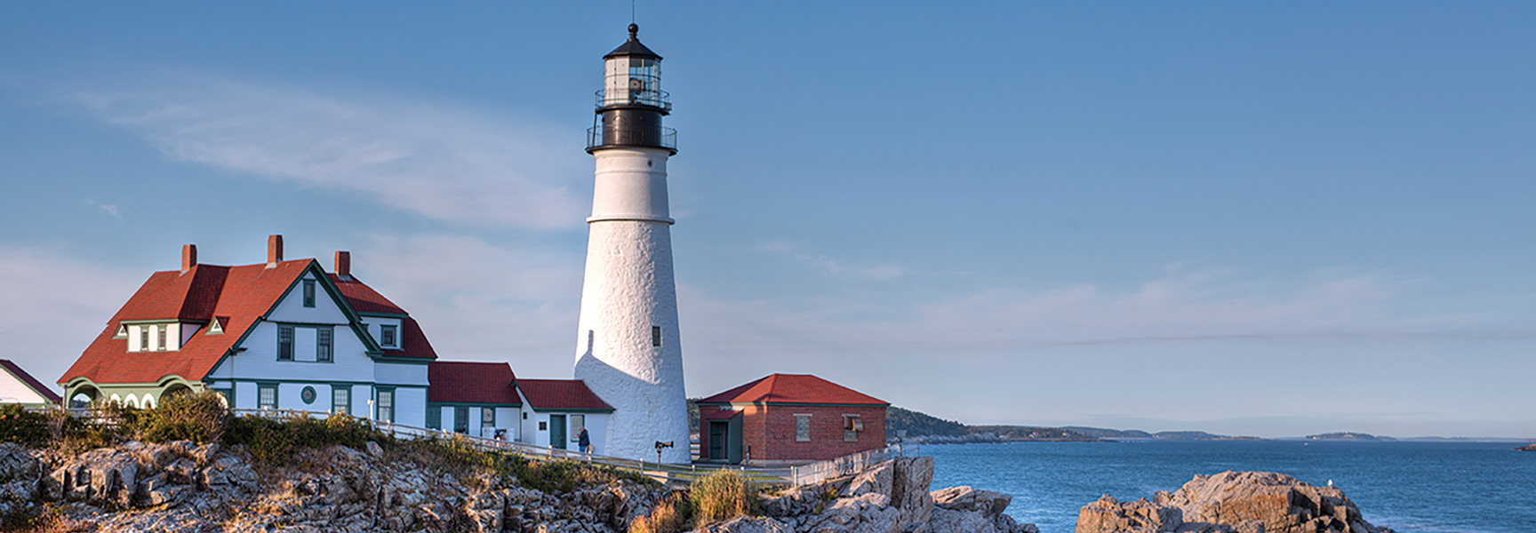 A classic white lighthouse and keeper's quarters overlook the ocean from the rocky coast of Maine on a sunny day.