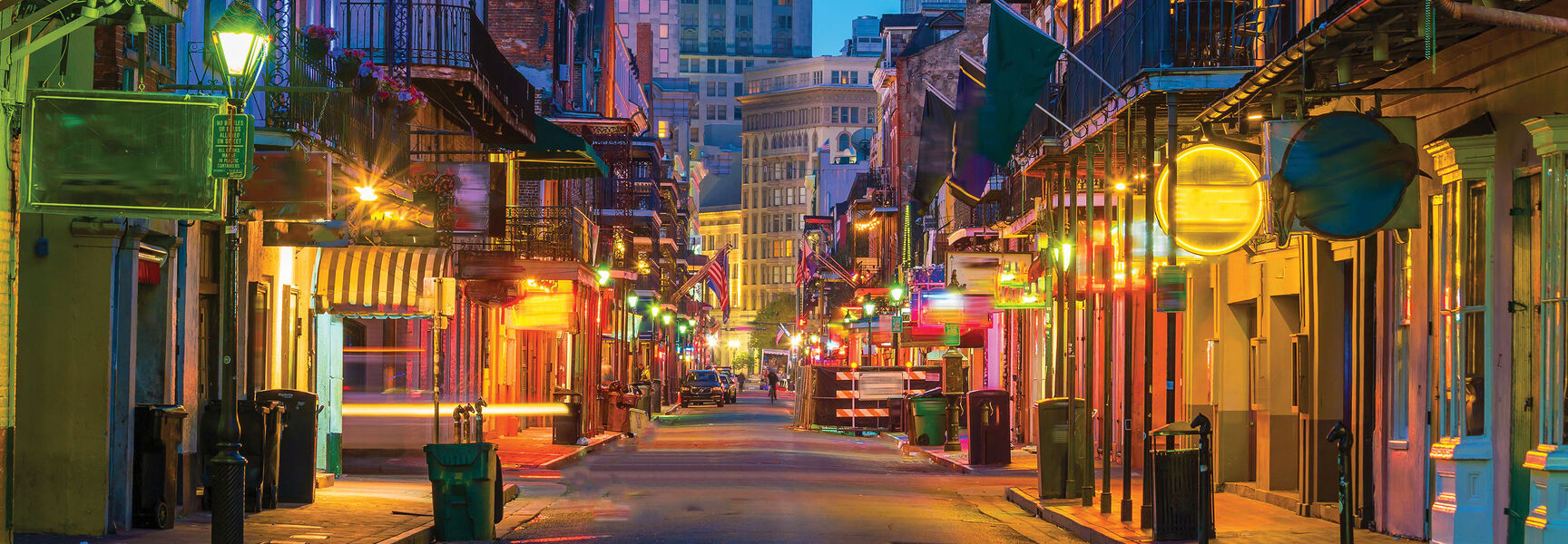 Vibrant twilight view of a street in the French Quarter of New Orleans featuring historic buildings with wrought-iron balconies and glowing neon signs.