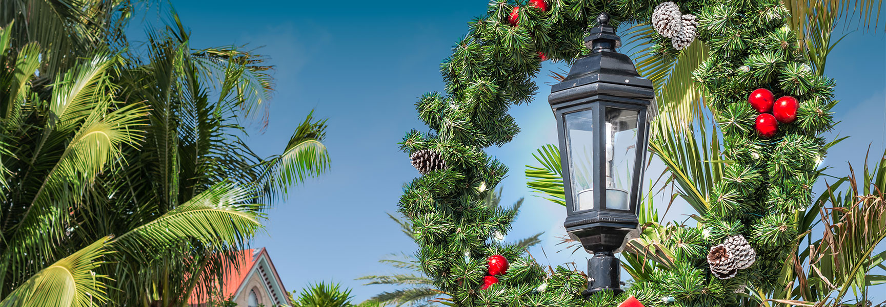 A black lamppost decorated with a festive Christmas wreath stands among palm trees against a sunny Florida sky.