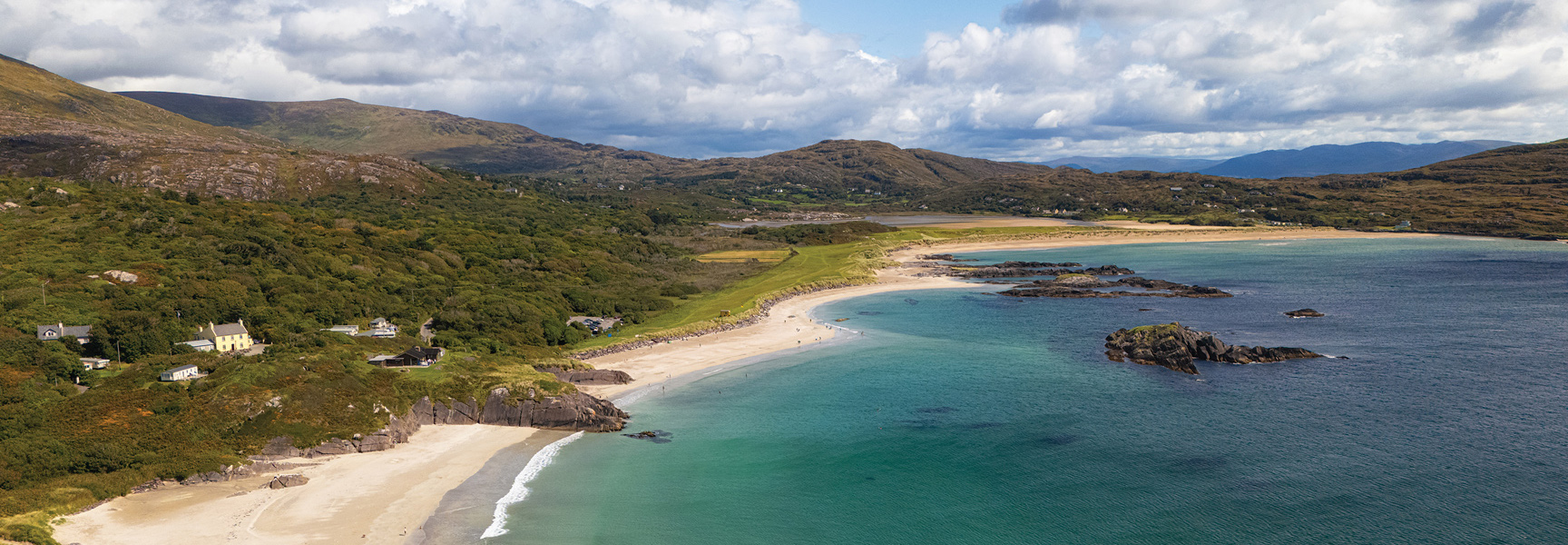 A sunny day on a sandy cove with turquoise water along the green, mountainous coast of Ireland under a partly cloudy sky.