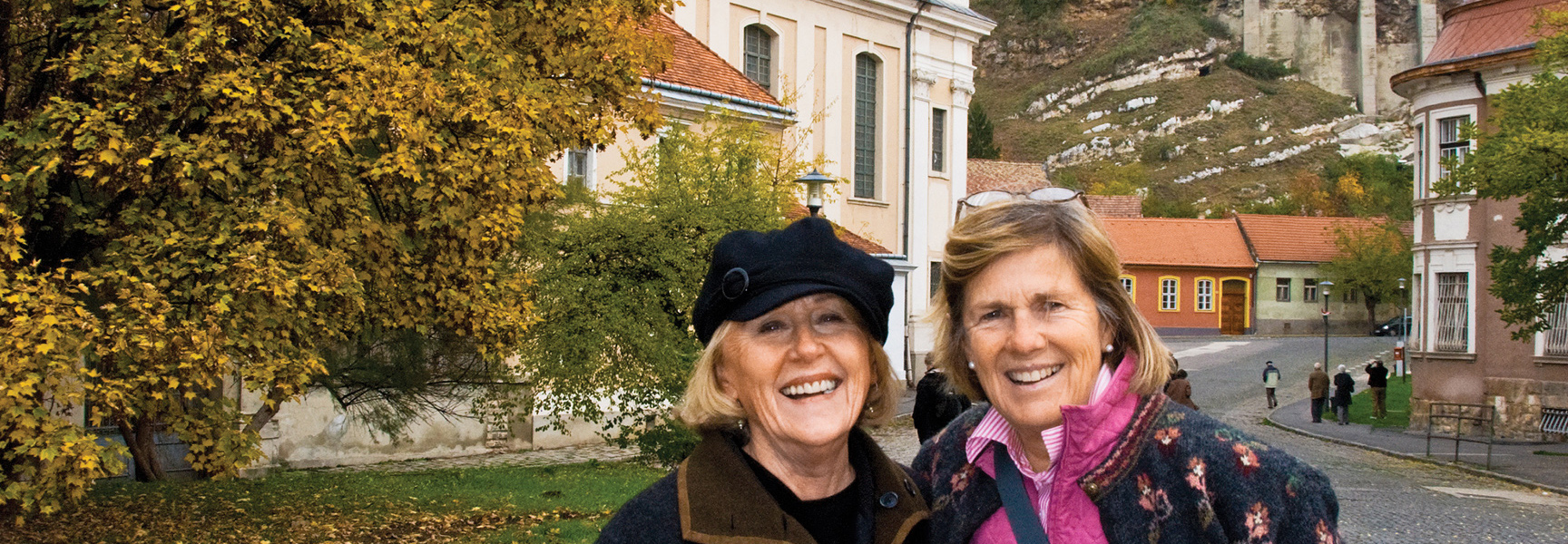 Two smiling women pose on a European street with autumn trees and historic buildings in the Czech Republic or Hungary.