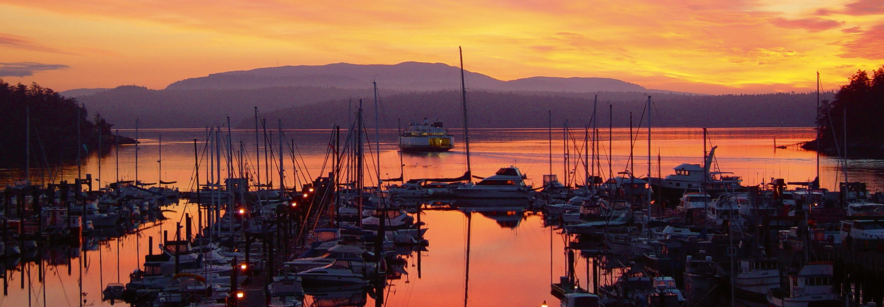 A colorful sunset reflects over a harbor full of boats in the San Juan Islands, Washington, as a ferry passes by distant mountains.