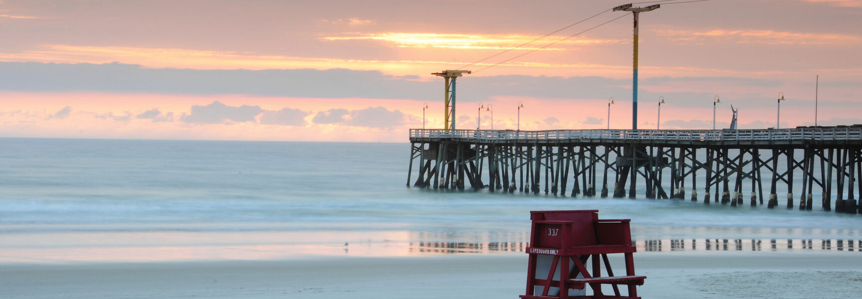 A red lifeguard stand on a sandy beach in Key West, Florida, with a wooden pier extending into the ocean under a pastel sunrise sky.