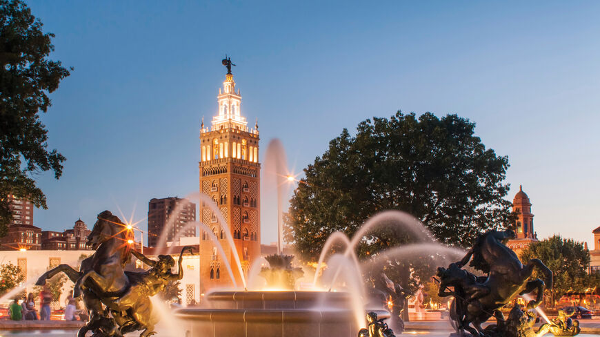 An illuminated fountain with bronze horse sculptures sprays water at twilight in Kansas City, Missouri, the Paris of the Plains, beneath an ornate tower.