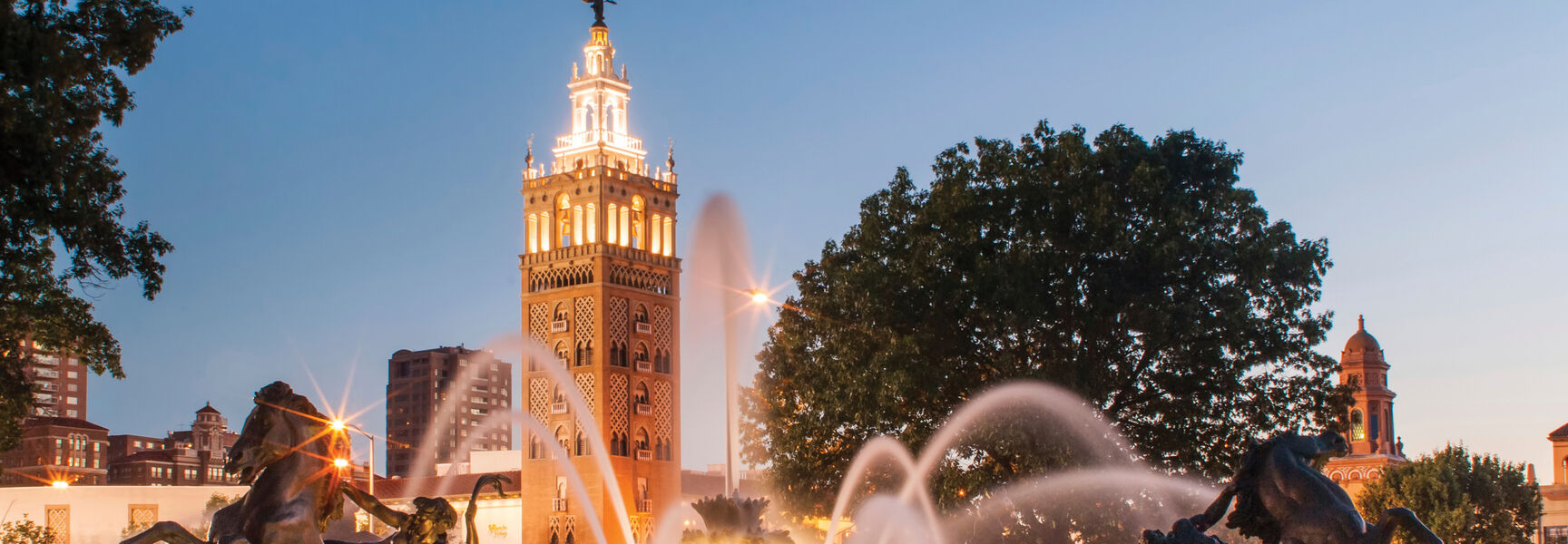 An illuminated fountain with bronze horse sculptures sprays water at twilight in Kansas City, Missouri, the Paris of the Plains, beneath an ornate tower.