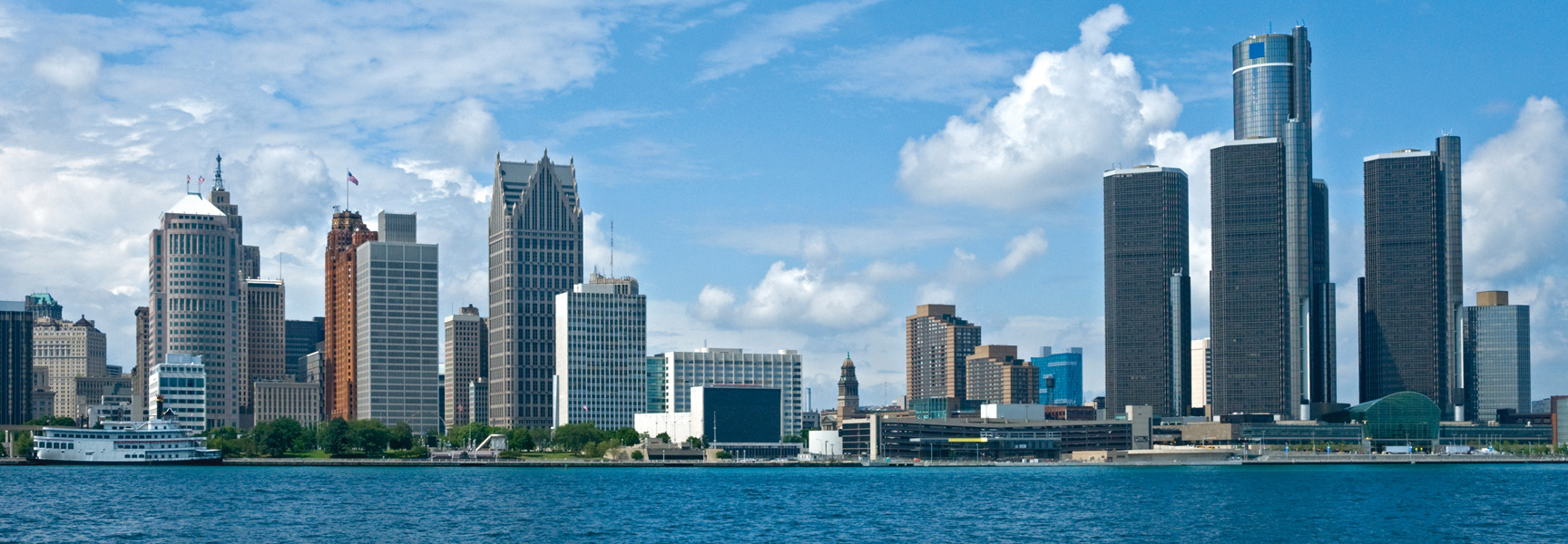 The Detroit, Michigan skyline, featuring the GM Renaissance Center, is viewed from across the water on a bright, partly cloudy day.