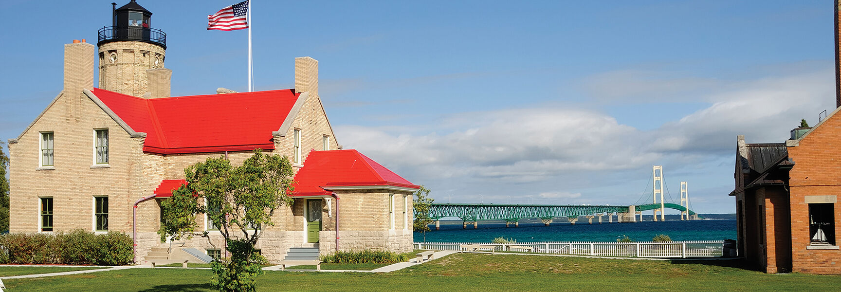 A historic brick lighthouse with a red roof and American flag stands before the Mackinac Bridge on the Great Lakes coast in Michigan.