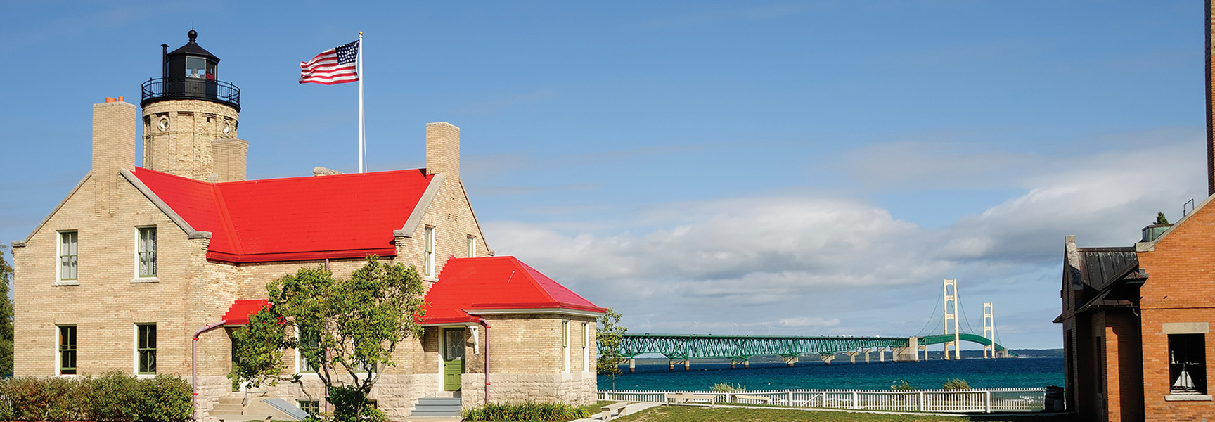 A brick lighthouse with a bright red roof overlooks the Mackinac Bridge spanning the blue waters of the Great Lakes.