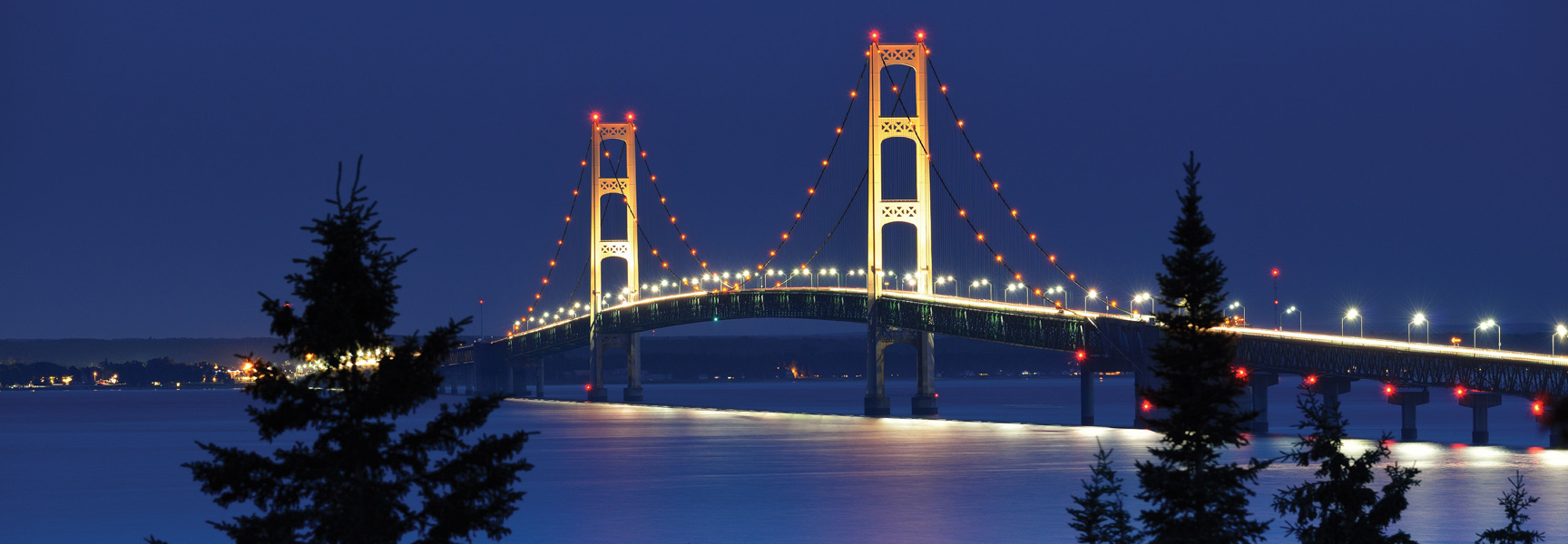 The Mackinac Bridge in Michigan is brightly illuminated at night, spanning the water with evergreen trees silhouetted in the foreground.