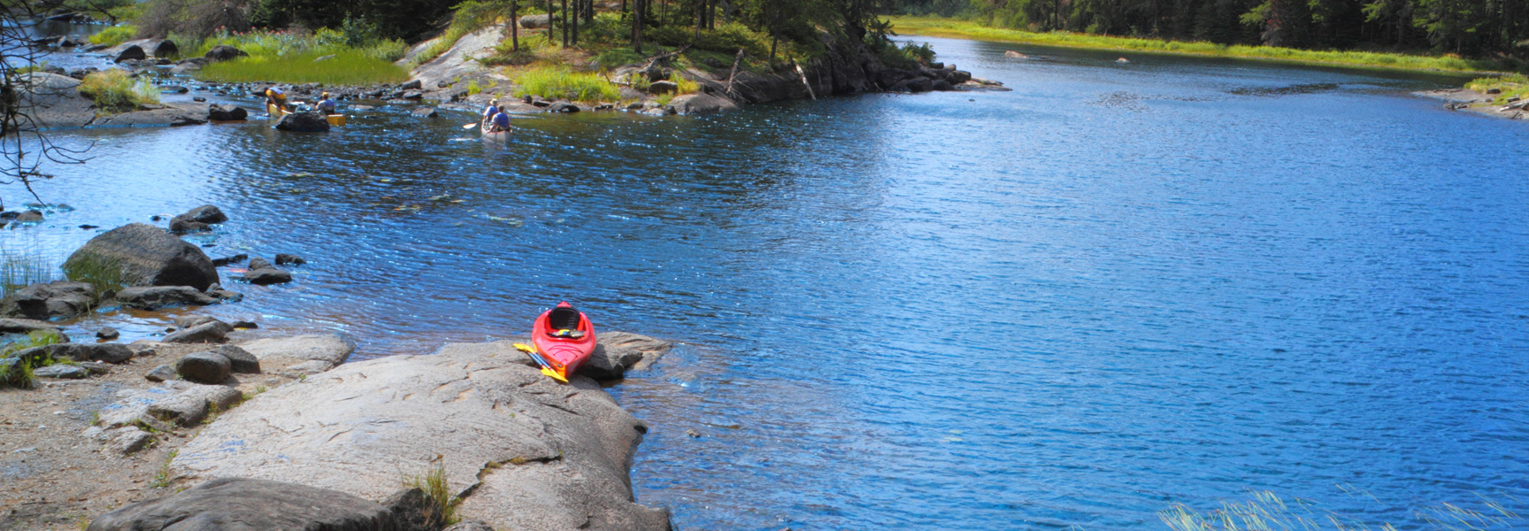 A red kayak sits on a rocky shore of a brilliant blue lake in Minnesota as people canoe in the background.