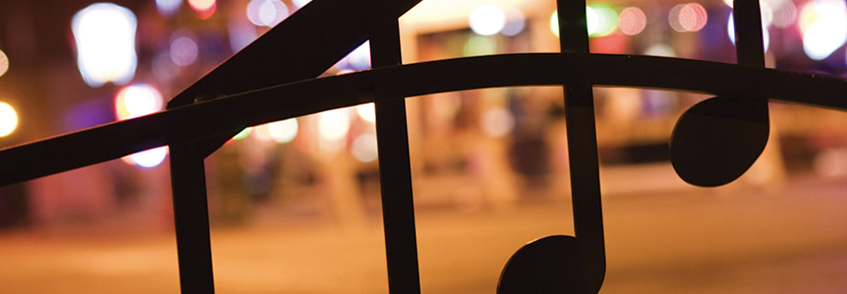 A close-up of a decorative iron gate with musical notes, with the blurred lights of a city street in Tennessee or Louisiana behind it.