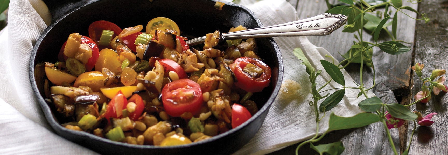 A rustic cast-iron skillet filled with Sicilian caponata, a mix of tomatoes, eggplant, and pine nuts, resting on a wooden table in Italy.