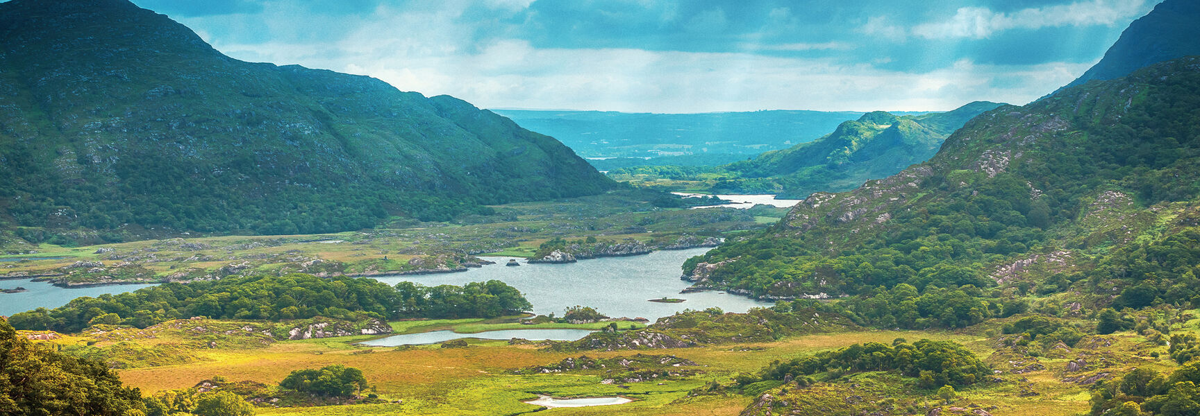 A scenic landscape of lush green mountains and lakes in the Emerald Isle of Ireland under a dramatic, cloudy sky.