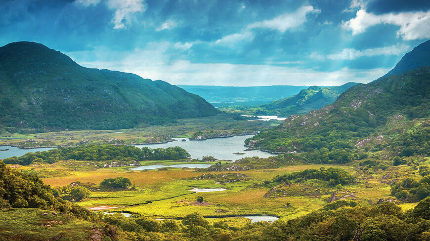 A scenic landscape of lush green mountains and lakes in the Emerald Isle of Ireland under a dramatic, cloudy sky.