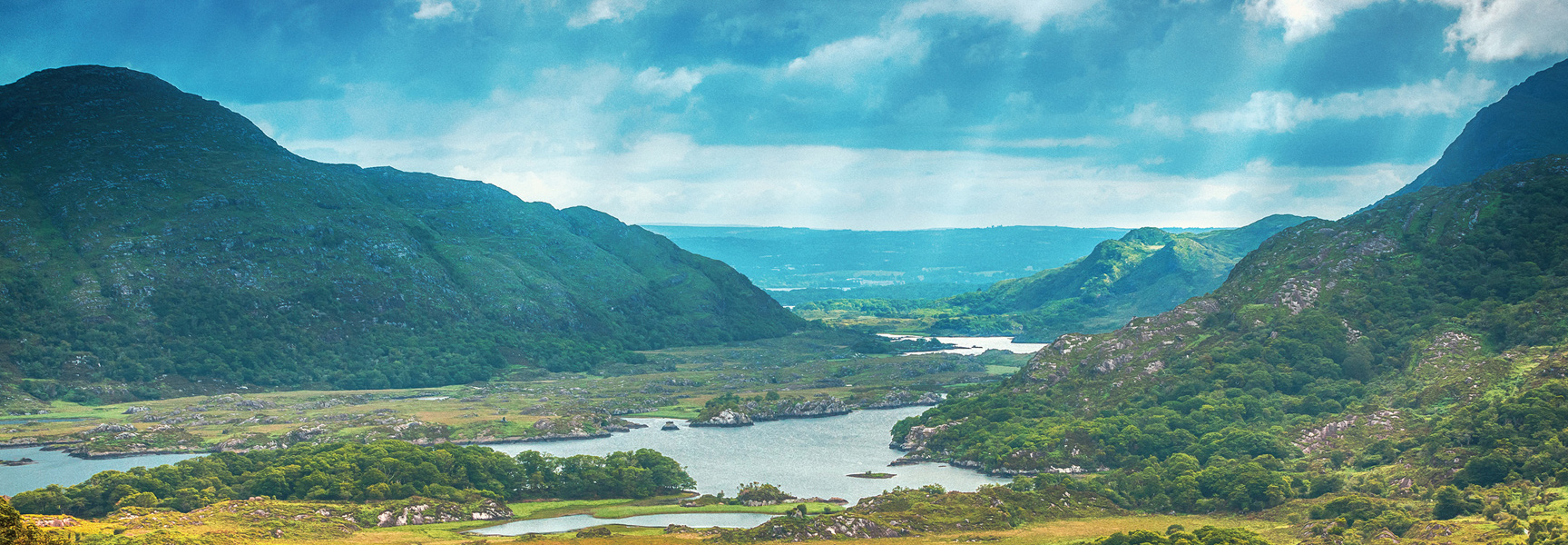 A scenic view of a lush green valley with mountains and lakes under a partly cloudy sky in Ireland.