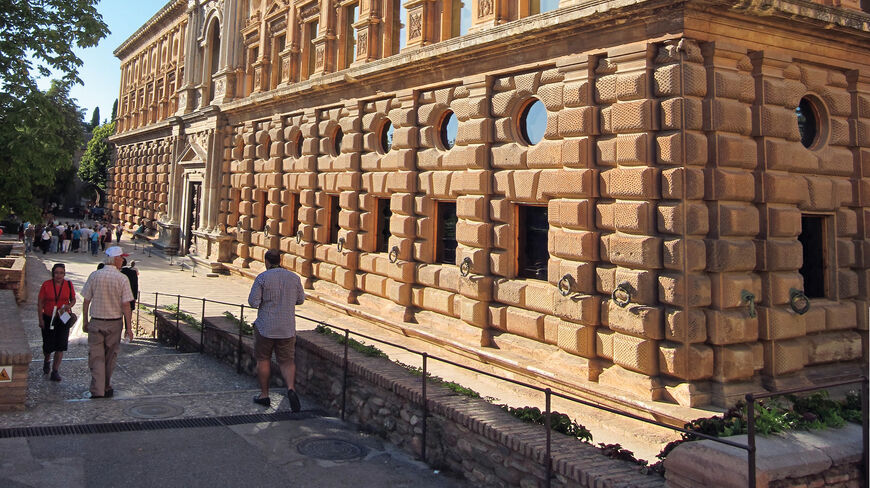 Tourists walk past the ornate, rusticated stone facade of the Palace of Charles V in Granada, Southern Spain.