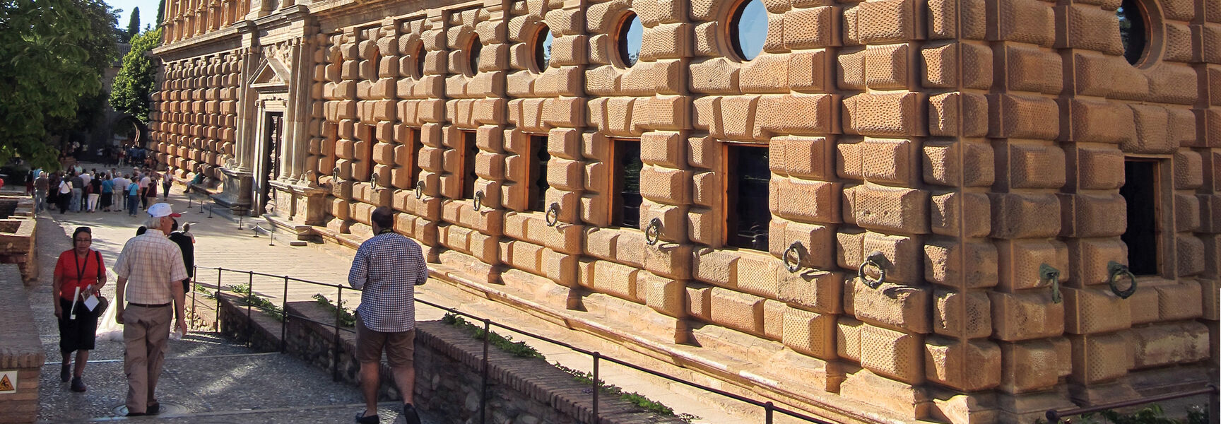 Tourists walk past the ornate, rusticated stone facade of the Palace of Charles V in Granada, Southern Spain.