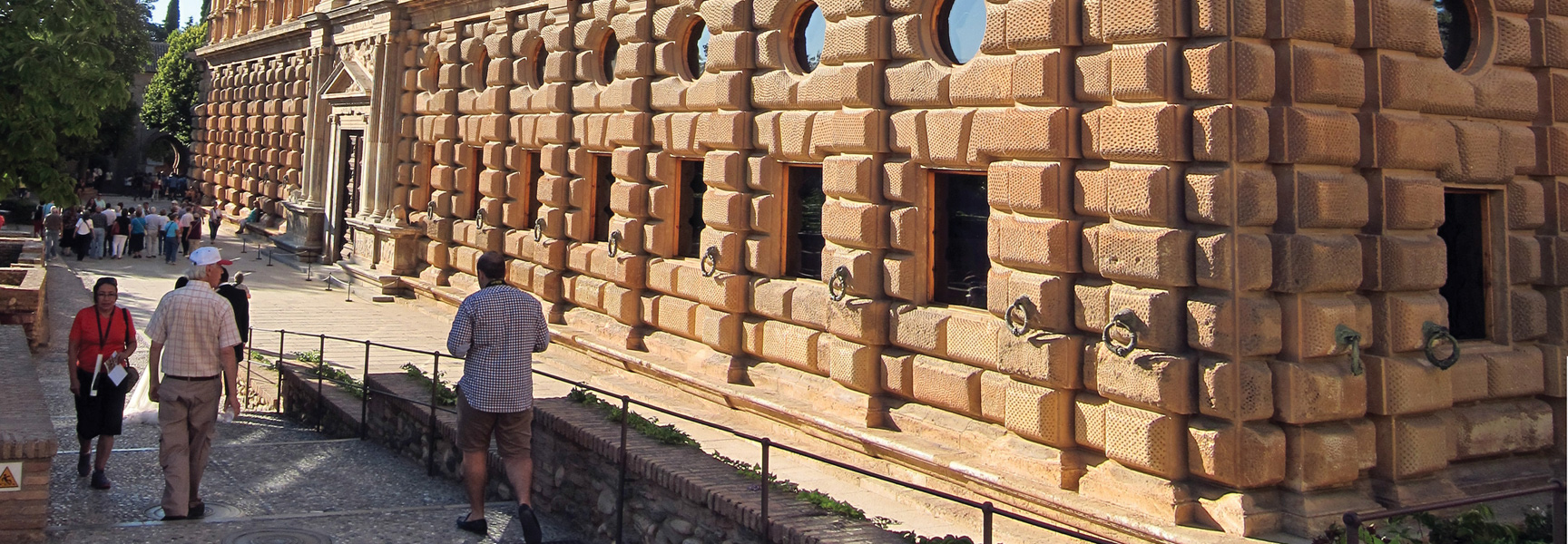 Tourists walk alongside the unique, textured stone exterior of a historic palace on a sunny day in Southern Spain.