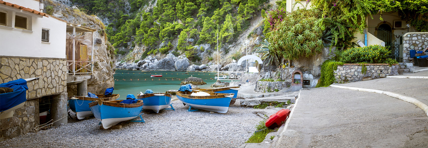 Blue boats rest on a pebble beach in a picturesque cove in Capri, Italy, with lush green cliffs in the background.