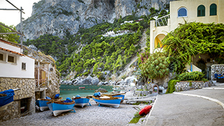 Small blue boats sit on a gravel beach in a cove in Capri, Italy, with buildings nestled into the tree-covered cliffs.
