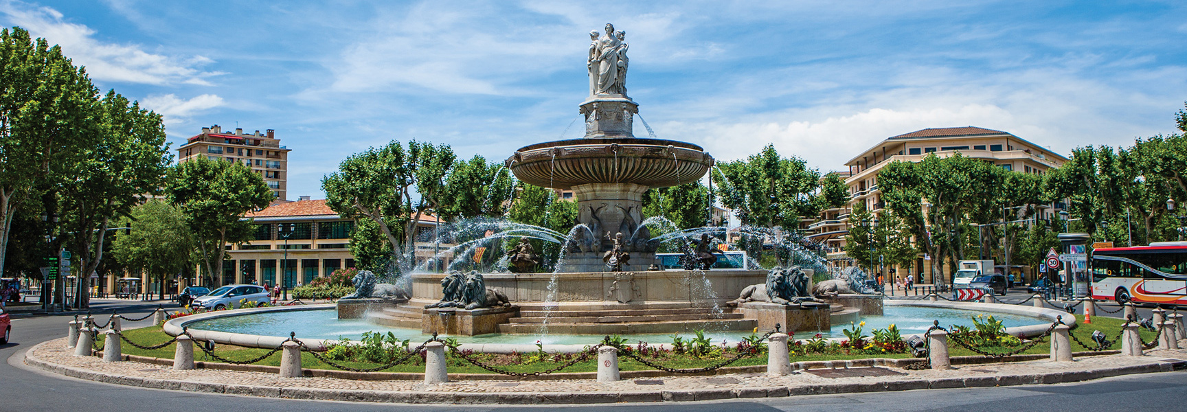 A large, ornate fountain with sculptures sits in the center of a traffic circle on a sunny day in Provence, France.