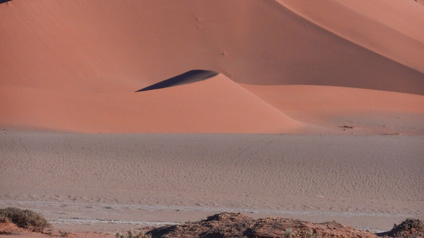 Towering red sand dunes with sharp ridges and deep shadows rise above scrubland in the Namib Desert of Namibia under a clear blue sky.
