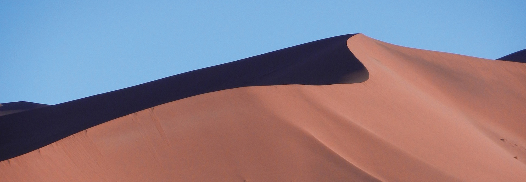 The curved crest of a red sand dune in Namibia creates a stark shadow against a clear blue sky.
