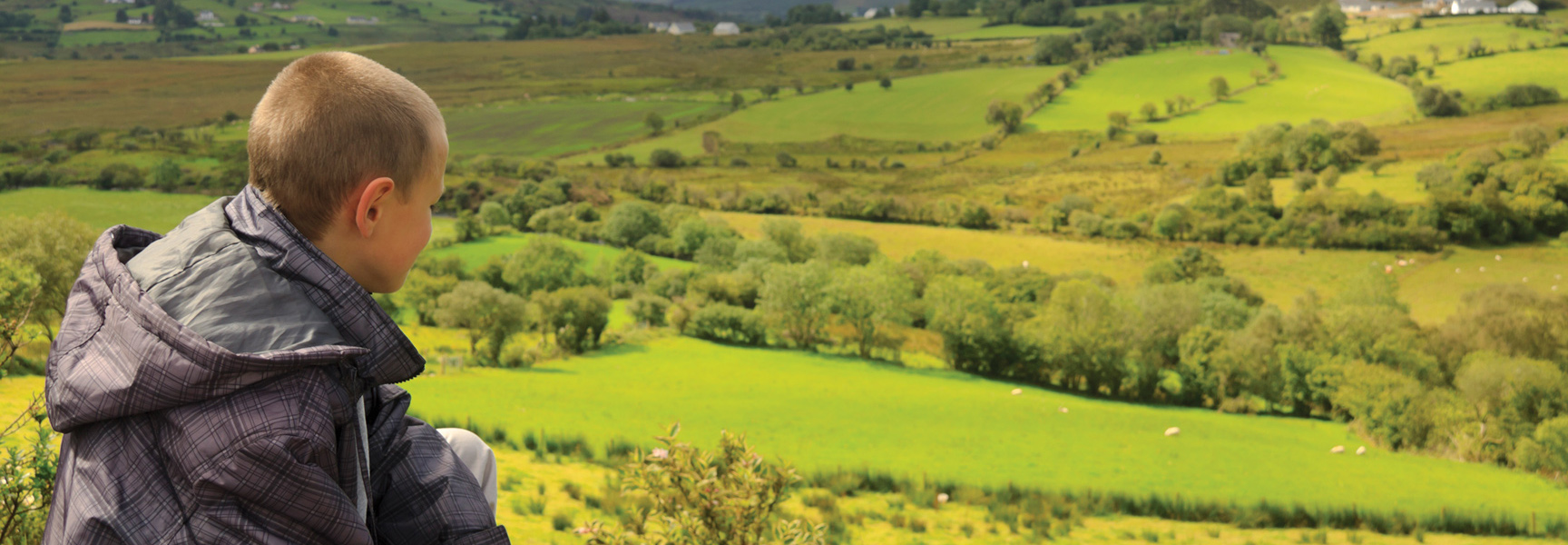A young boy looks out over a vast landscape of rolling green hills and fields in the Irish countryside.