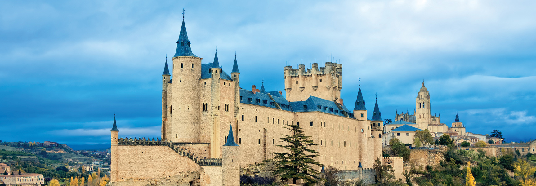 The Alcázar of Segovia, a historic stone castle with distinctive blue turret roofs, sits on a rocky crag in Spain.