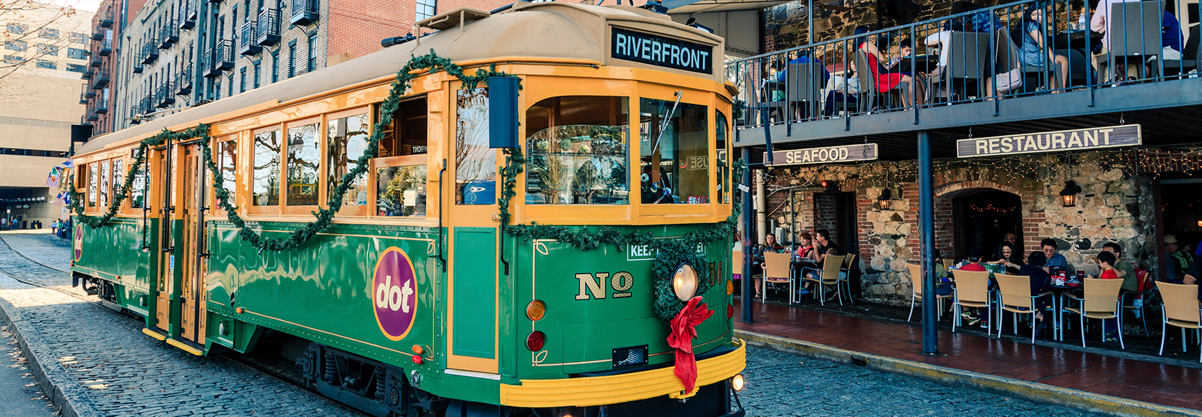 A green and yellow holiday trolley decorated with garlands drives down a cobblestone street in Savannah, Georgia next to a seafood restaurant.