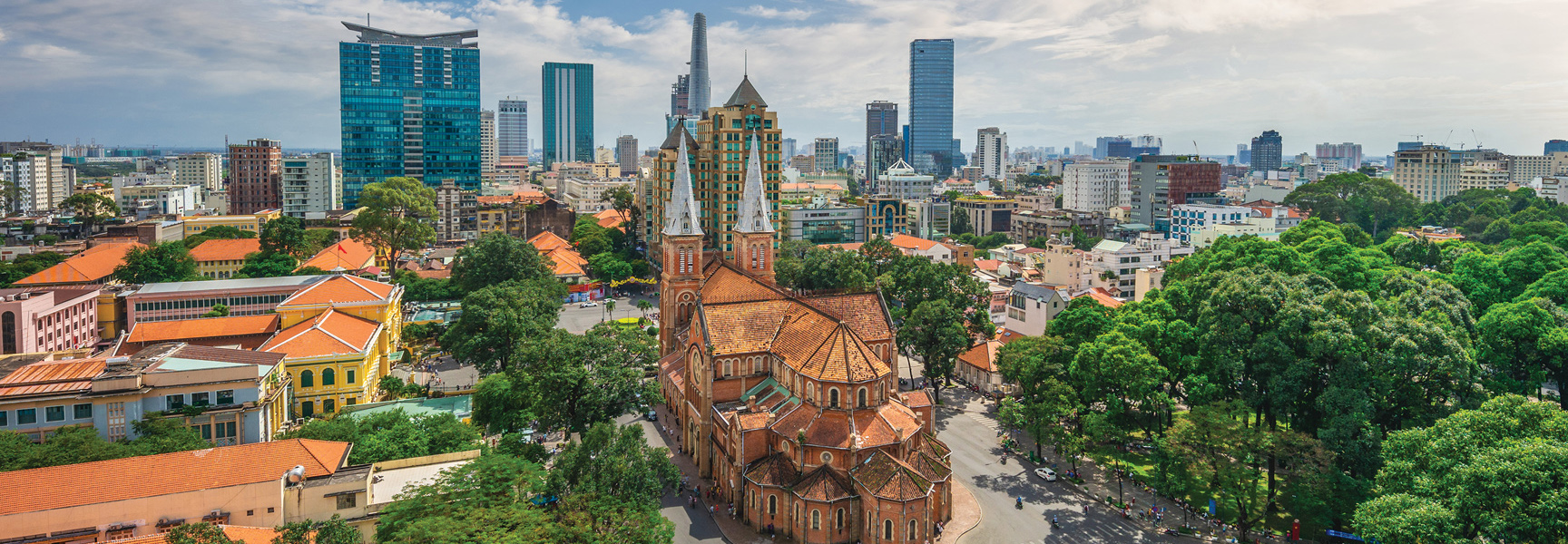 An aerial view of the Saigon Notre-Dame Cathedral surrounded by lush trees and the modern Ho Chi Minh City, Vietnam skyline under a partly cloudy sky.