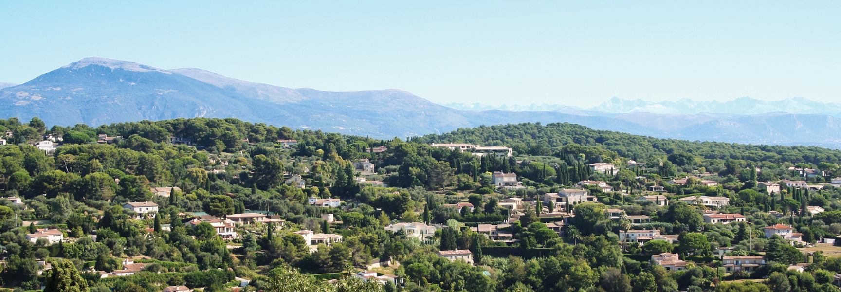 A scenic view of houses nestled in the green hills of the French Riviera with mountains in the background.