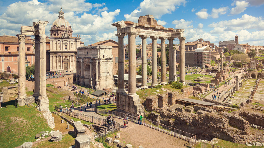 Ancient ruins of the Roman Forum in Rome, Italy, featuring stone columns and historical architecture under a bright, cloud-filled blue sky.