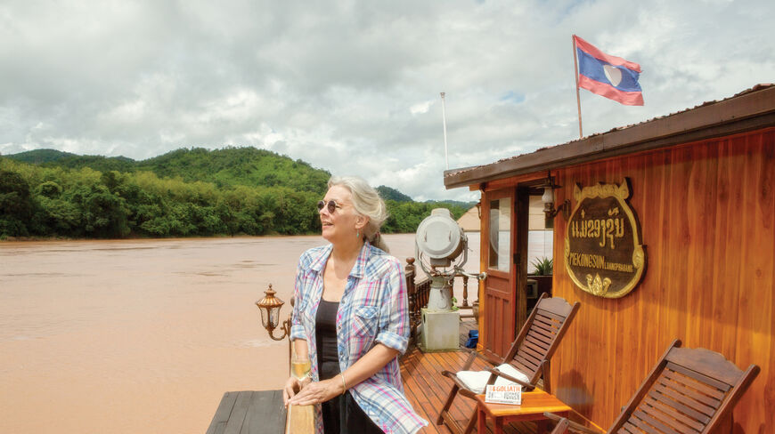 A woman stands on the wooden deck of a boat on the Mekong River in Luang Prabang, Laos, overlooking lush green hills.