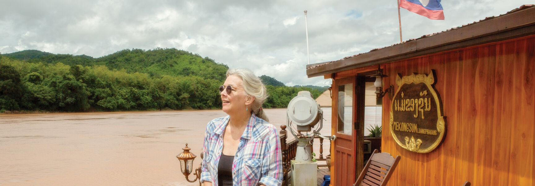 A woman stands on the wooden deck of a boat on the Mekong River in Luang Prabang, Laos, overlooking lush green hills.