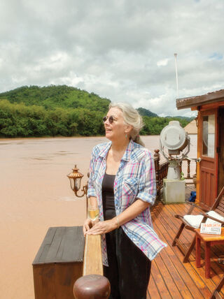 A woman stands on the wooden deck of a boat on the Mekong River in Luang Prabang, Laos, overlooking lush green hills.