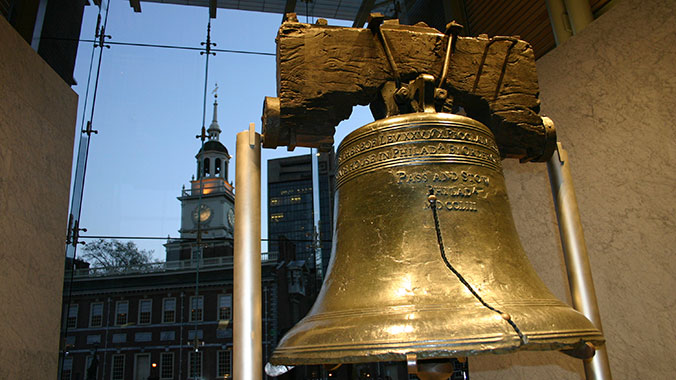 A close-up of the Philadelphia Liberty Bell with its iconic crack, displayed indoors before a large window overlooking Independence Hall at dusk.