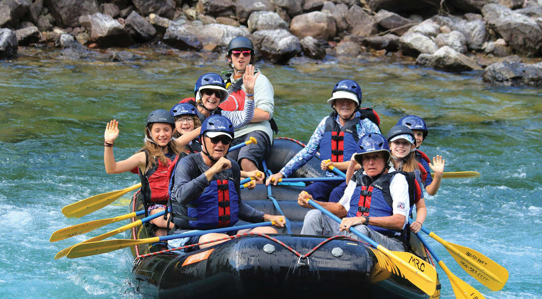 A family wearing helmets and life jackets smiles and waves while rafting on the turquoise Flathead River in Montana’s Glacier National Park.