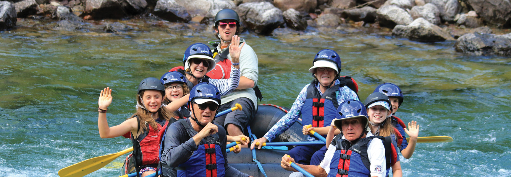 A multi-generational family in helmets and life vests smiles and waves while rafting down a river in Glacier National Park, Montana.