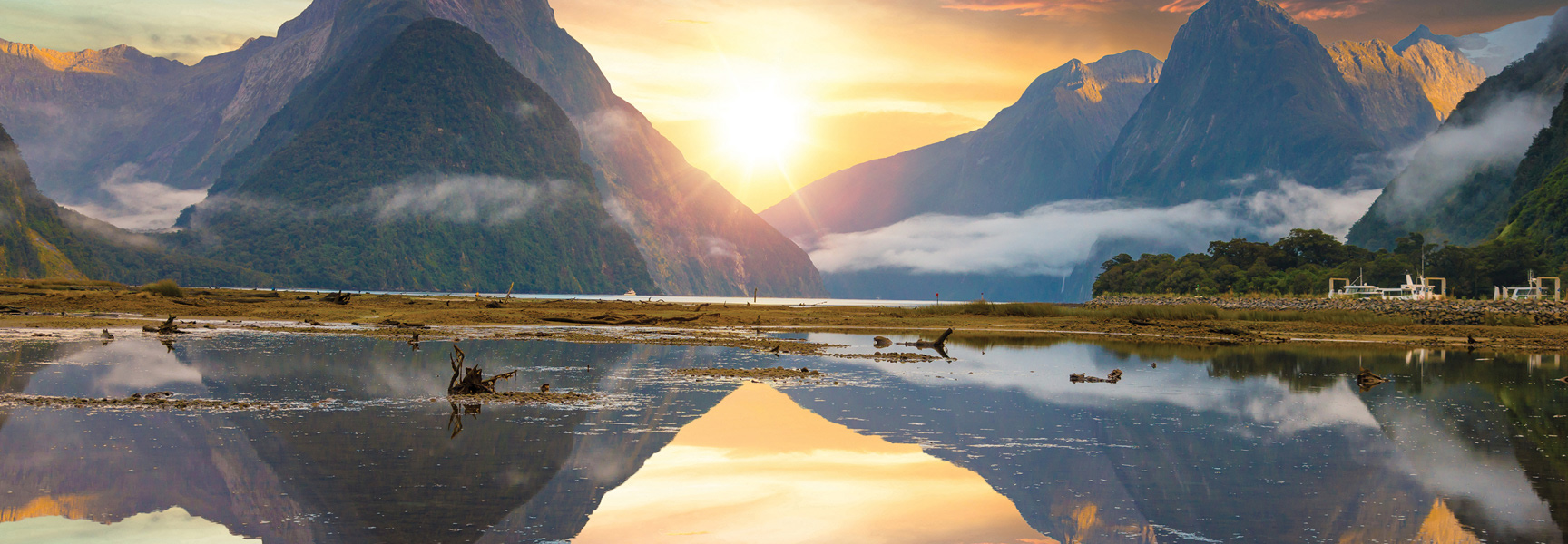 A golden sunset reflects on the water of a mountainous fjord in New Zealand.