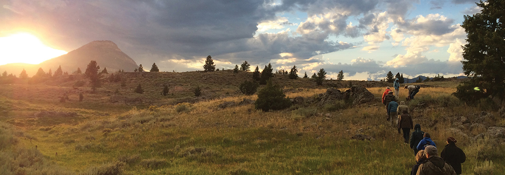 A group of people hike through a grassy, mountainous landscape in Wyoming at sunrise, with a prominent peak visible under a partly cloudy sky.
