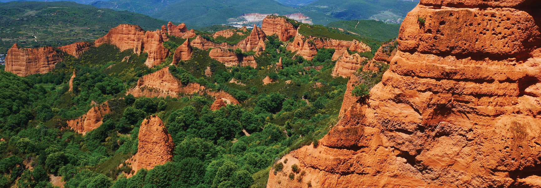 Jagged red rock formations covered with lush green trees rise towards a mountain range in Spain and Portugal.