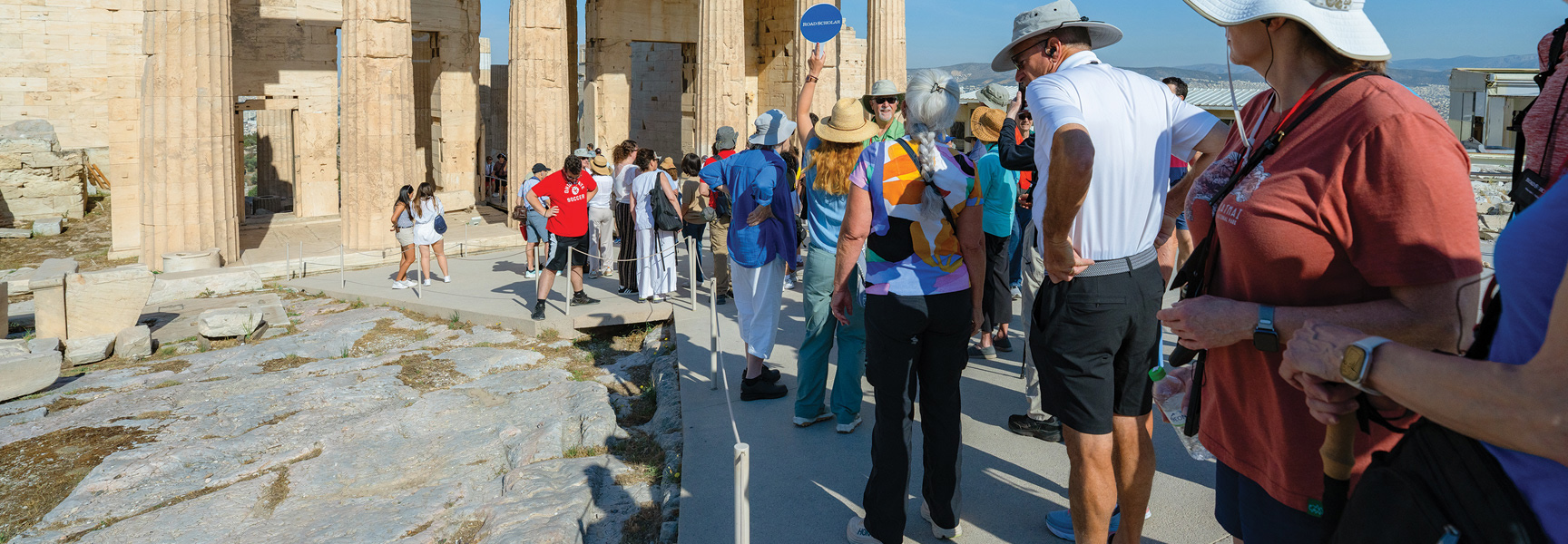 A tour group explores the ancient stone ruins of the Acropolis in Greece on a sunny day.