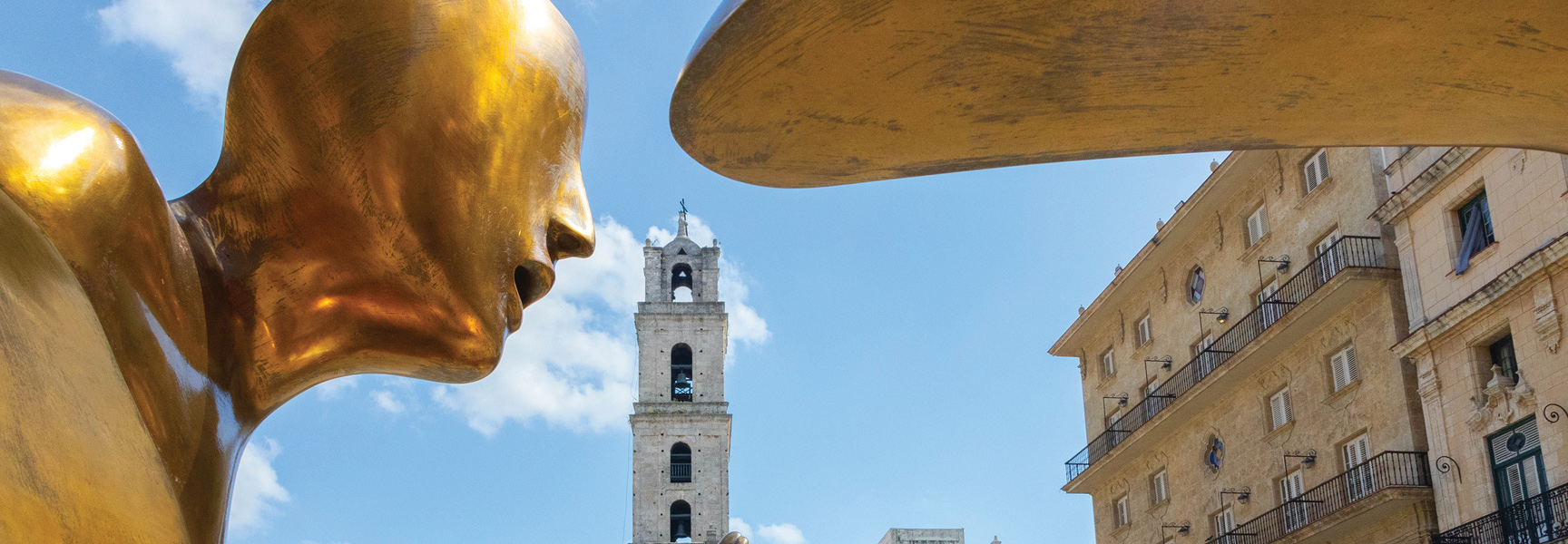 A large, golden sculpture of a face in profile stands before the historic bell tower of the Basilica Menor de San Francisco de Asís in Havana, Cuba.