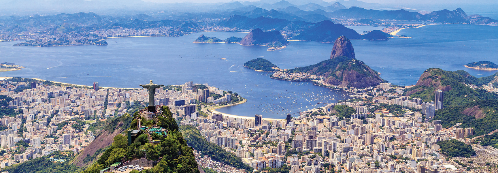An aerial view of Rio de Janeiro, Brazil, with the Christ the Redeemer statue overlooking the city, Sugarloaf Mountain, and Guanabara Bay's blue water.