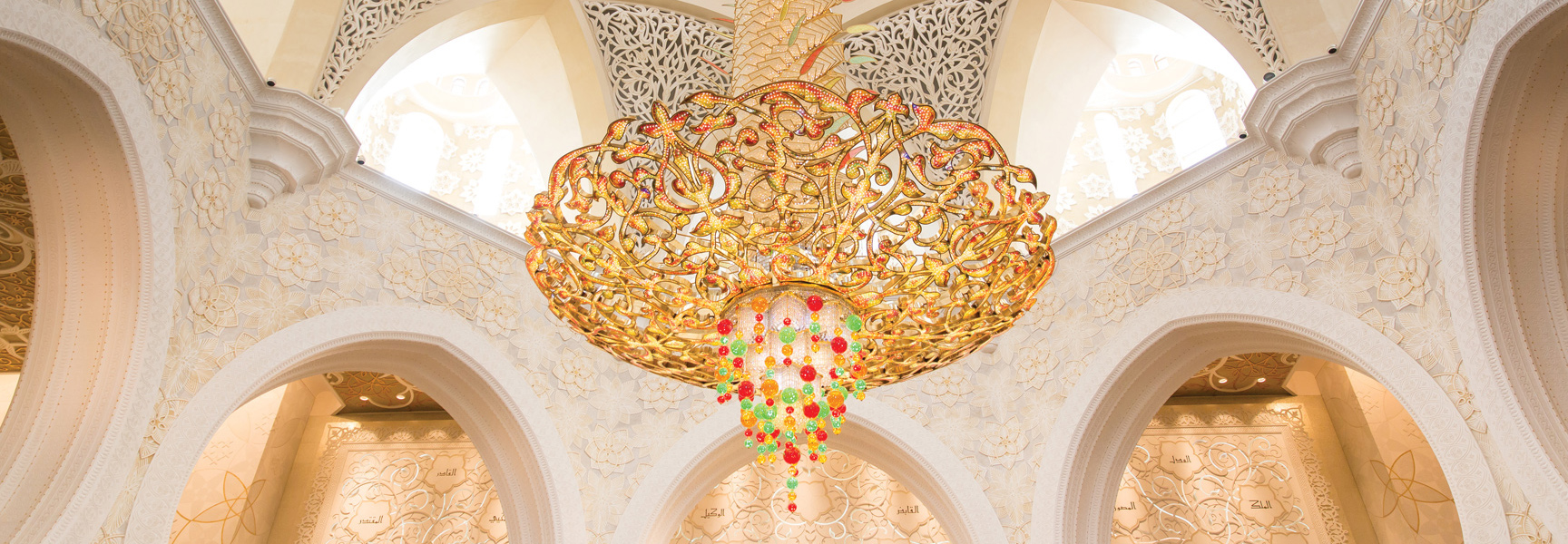 An ornate, golden chandelier with colorful glass details hangs from the ceiling of a grand hall with intricately carved white arches and walls.