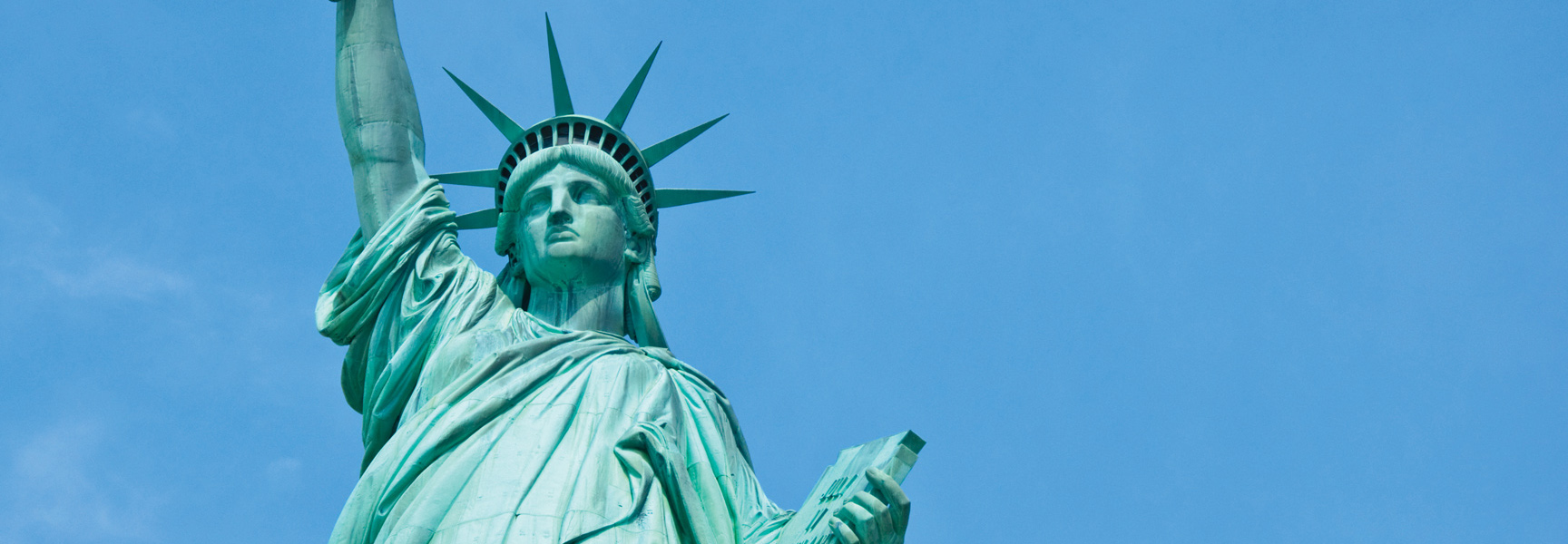 A close-up of the Statue of Liberty in New York, looking up at her face and raised torch against a bright blue sky.