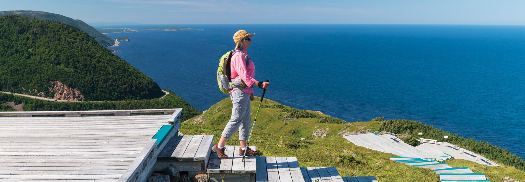 A woman with a backpack and hiking poles enjoys the view of the ocean from a wooden boardwalk on the coast of Nova Scotia.