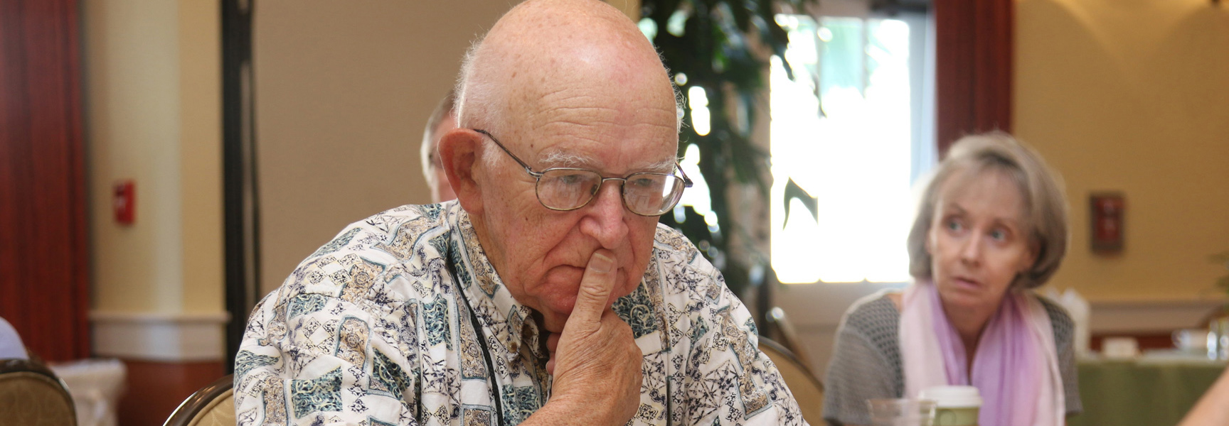 An older man with glasses concentrates during a bridge lesson in Vermont, with a woman looking on in the background.